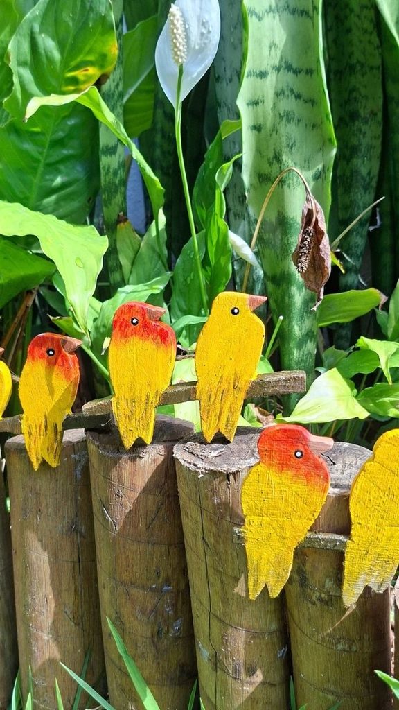 Passarinhos de madeira pintados em amarelo e laranja sobre cerca de toras, com plantas ao fundo