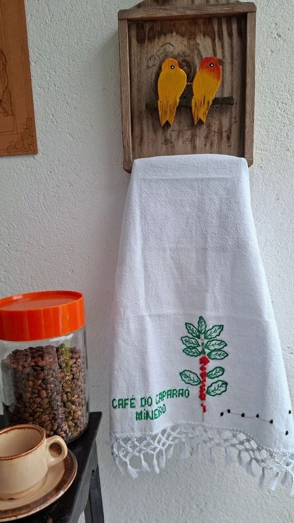 Pano de prato branco bordado com ramo de café e a frase Café do Caparaó Mineiro, pendurado em porta-pano de madeira com passarinhos pintados
